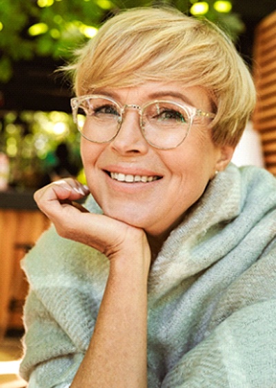 A middle-aged woman wearing glasses and a sweater, smiling after receiving implant dentures in Westminster