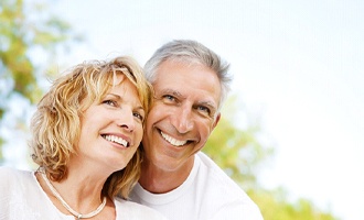 A middle-aged couple, both wearing white and smiling while standing outdoors, pleased with their new teeth