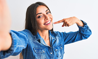 A young woman wearing a denim button-down shirt and pointing to her whiter, brighter smile