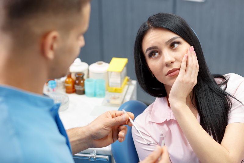 a young woman holding her cheek in pain while listening to a dentist explain the need for a root canal